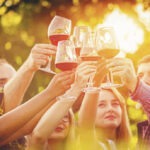 Multiracial group of young friends toasting wine glasses at outdoor party - Friendship celebration with happy people enjoying time together at vineyard restaurant garden drinking red wine at sunset